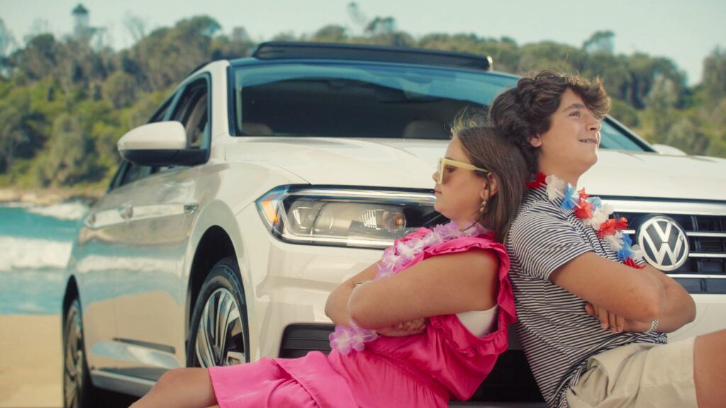 Two kids relax on the beach in front of a brand new car
