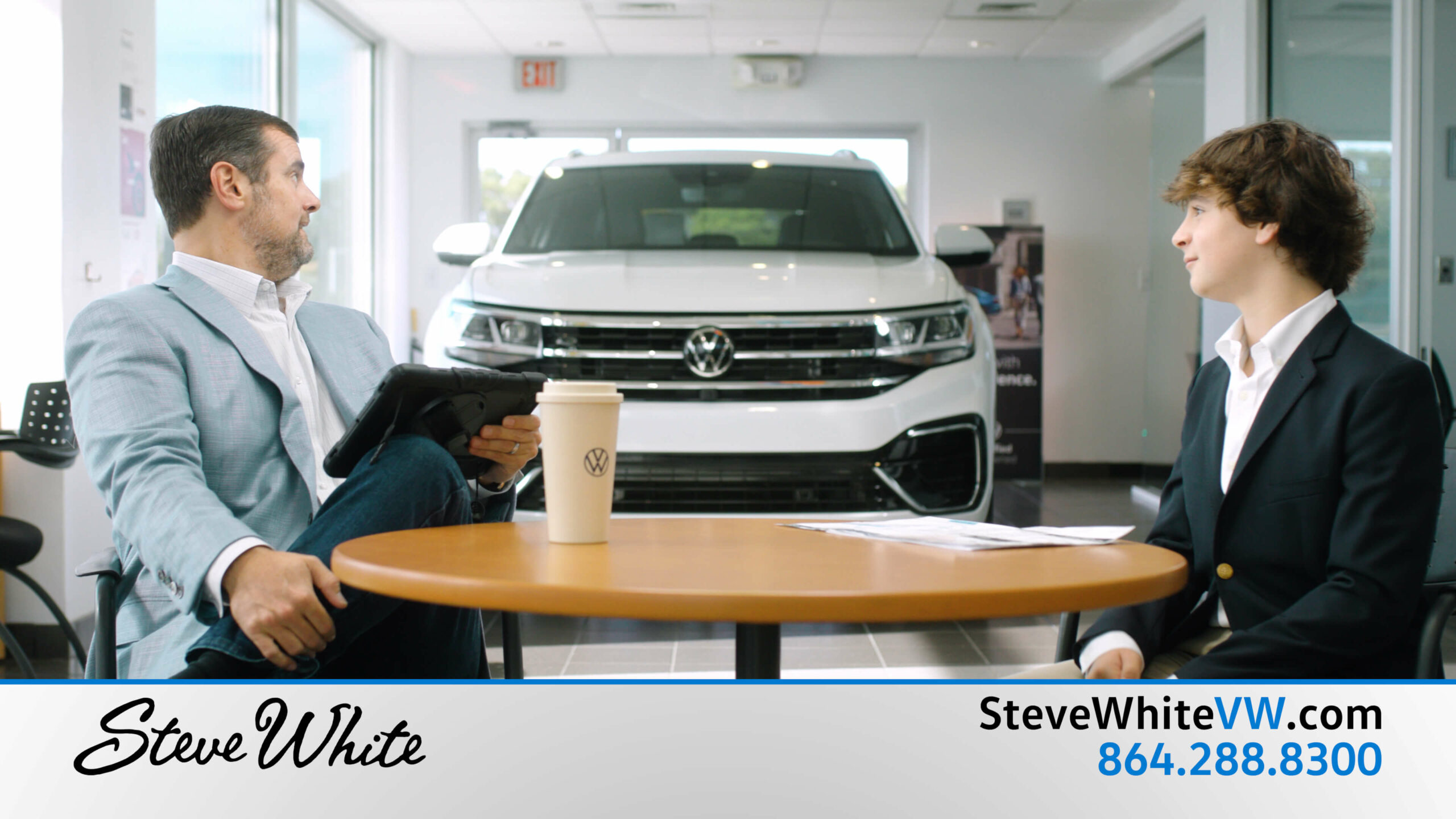 A car dealer owner and his son sit at a round table. a white volkswagen is parked in the background.